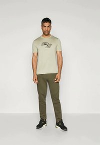 Vīrietis stāv pretī, tērpies gaiši zaļā grafiskā T-kreklā, olīvzaļās biksēs un melnās sporta kurpēs ar zaļiem akcentiem.