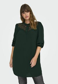 Tumšo zaļo tuniku ar mežģīņu paneļiem uz krūtīm, kas tiek piedāvāta ar pogājamu kakla izgriezumu un puffām piedurknēm. Audums izskatās gluds un viegls.