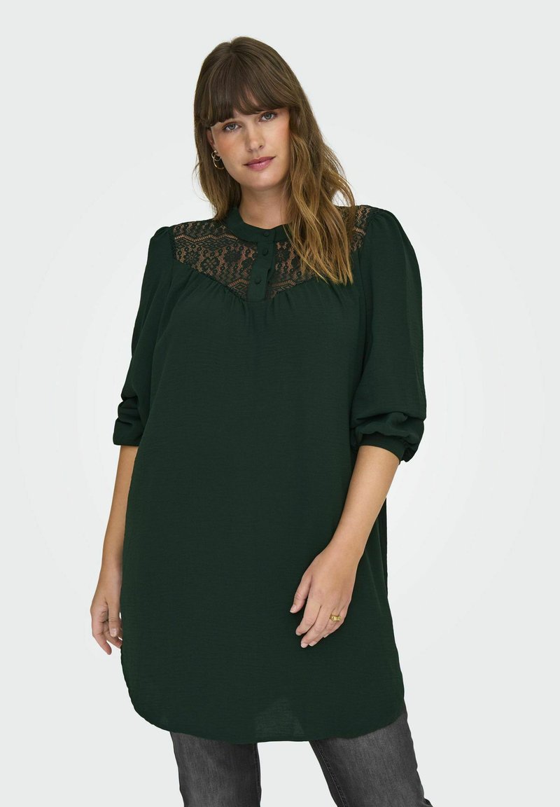 Tumšo zaļo tuniku ar mežģīņu paneļiem uz krūtīm, kas tiek piedāvāta ar pogājamu kakla izgriezumu un puffām piedurknēm. Audums izskatās gluds un viegls.