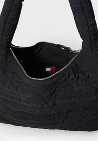Sac fourre-tout en tissu matelassé noir avec deux poignées, présentant une marque texturée, une fermeture éclair et une poche intérieure pour le rangement.