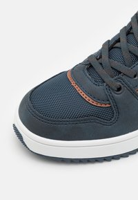 Mørkeblå idrettssko med strukturert mesh-materiale, brune imitert lærdetaljer, grå snører og en hvit gummisåle med godt feste.