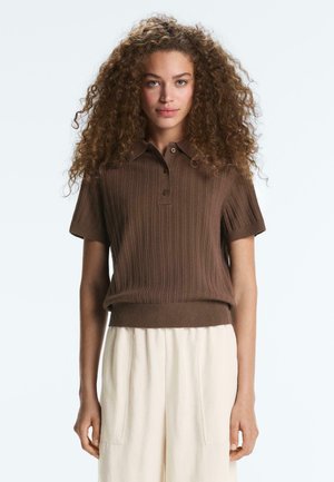 Žena s kučeravými vlasmi, ktorá nosí hnedé pletené polo s krátkym rukávom a krémové nohavice s vysokým pásom, stojí na jednofarebnom pozadí.