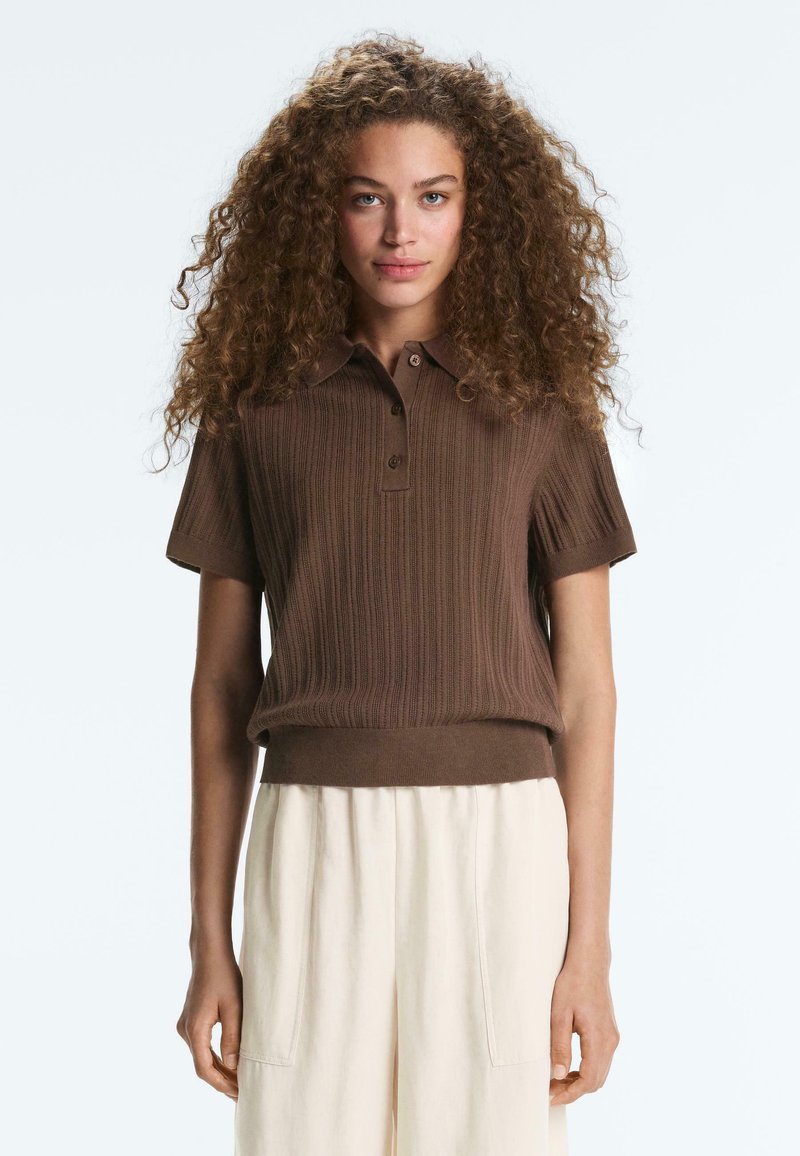 Femme aux cheveux bouclés portant un polo en maille marron à manches courtes et un pantalon crème taille haute, debout devant un fond uni.