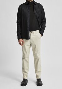 Černá košile na knoflíky přes černý rolák, spárovaná se světlými krémovými kalhotami a černými botami. Hladká tkanina, přiléhavý střih, jednoduchý design.