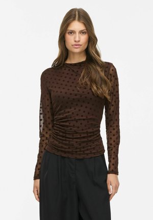 Femme portant un haut marron transparent à manches longues avec des pois et des côtés froncés, associé à un pantalon noir taille haute.