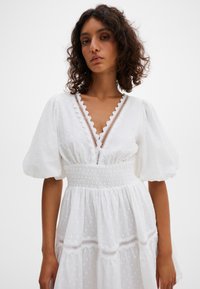 Moteris su garbanotais plaukais, vilkinti baltą suknelę su pūstomis rankovėmis, V formos iškirpte, nėrinių apdaila ir elastinga juosmens dalimi, stovinti priešais paprastą foną.