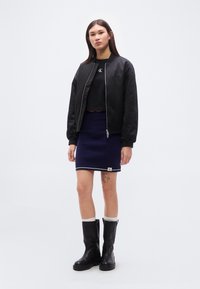 Μαύρο μπουφάν bomber, εφαρμοστό μαύρο crop top, υφασμάτινη μπλε φούστα με λευκή επένδυση, μαύρες μπότες μέχρι το γόνατο, το μοντέλο στέκεται μπροστά σε απλό φόντο.