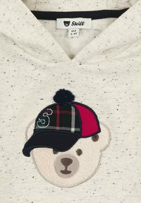 Elfenbeinfarbener Hoodie mit einem Bärenkopf-Design, das einen strukturierten, hellbraunen Fellfleck zeigt, sowie einer mehrfarbigen Mütze mit Bommel und dem Buchstaben "S".
