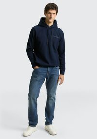 Marineblauer Kapuzenpullover mit Kordelzug, gerippten Bündchen und kleinem Logo auf der Brust, kombiniert mit hellblauen Jeans und weißen Turnschuhen.