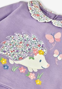 Felpa viola con ricamo di un riccio e stampa floreale sulla schiena, circondato da fiori colorati e farfalle. Collo a scallop.