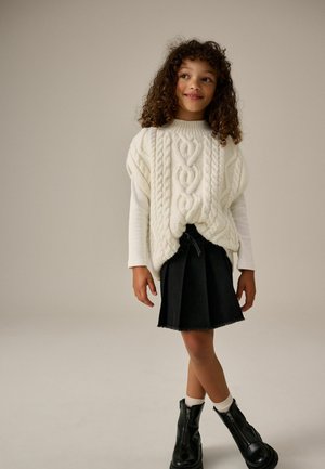 Pull en tricot torsadé crème sur un haut à manches longues blanc, associé à une jupe plissée noire et des bottines noires à zips.