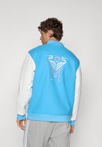 Light blue jacket with white sleeves, featuring a colorful graphic of herons and a sun on the back. Soft texture, ribbed cuffs.