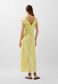 Long dress with a V-back, cap sleeves, and a watercolor pattern in green and yellow tones. Soft fabric with a flowing design. Black strappy sandals.