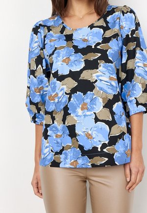 Frau trägt eine schwarze Bluse mit großem blauem Blumenmuster und Dreiviertelärmeln, kombiniert mit beigen Hosen.