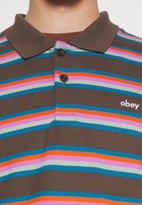 Polo marron avec des rayures horizontales en bleu, orange, rose et vert. Patte de boutonnage à deux boutons et petit logo brodé sur la poitrine.