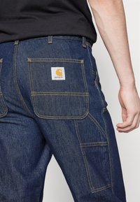 Denim cargo pants in dark blue with yellow stitching. Features two back pockets and a square logo patch on the right pocket.