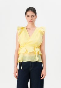 Mujer vistiendo una blusa amarilla sin mangas, transparente y con volantes, combinada con pantalones oscuros de tiro alto, sobre un fondo blanco liso.