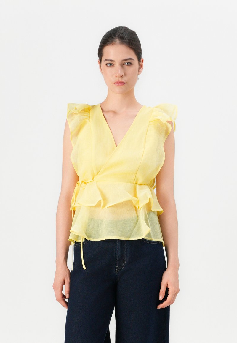 Mujer vistiendo una blusa amarilla sin mangas, transparente y con volantes, combinada con pantalones oscuros de tiro alto, sobre un fondo blanco liso.