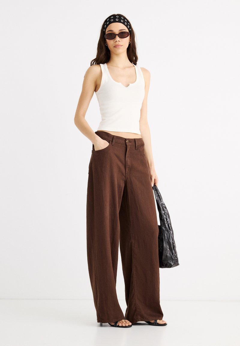 Woman wearing white sleeveless top, wide brown pants, black headband with white circles, sunglasses, holding black textured bag.