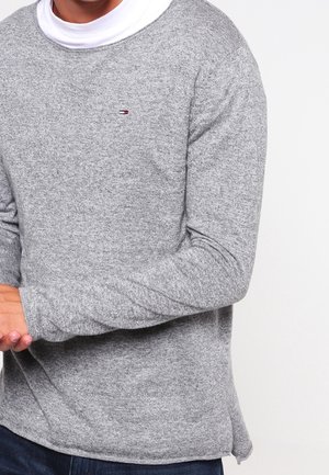 Mann trägt einen grauen Pullover mit langen Ärmeln über einem weißen Hemd, mit vor dem Körper verschränkten Händen und sichtbar dunkelblauer Hose.