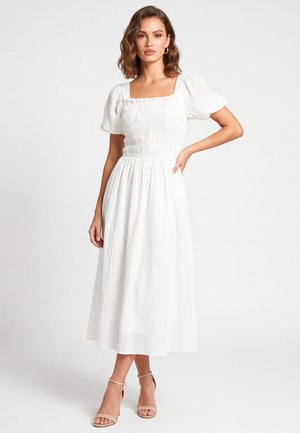 Donna con un vestito midi bianco con maniche a sbuffo e corpetto arricciato, in piedi con una mano sulla vita e sandali beige con tacco alto.