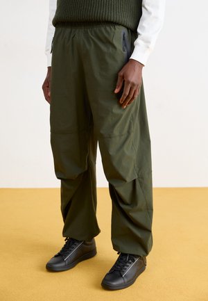 Pantaloni cargo verzi, ușori, cu talie elastică, având buzunare cu fermoar și o croială lejeră. Asortați cu adidași negri, de tip high-top.