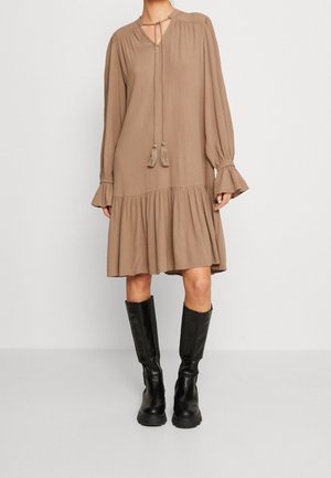 Kvinde iført en løs, langærmet, knælængde beige kjole med kvastbind og sorte knæhøje støvler, stående foran en ensfarvet baggrund.