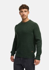 Suéter de punto acanalado verde oscuro con cuello redondo, mangas largas y un pequeño parche de marca en el dobladillo. Textura suave, ajuste casual.