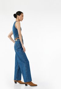 Denim crop-top ja leveät farkut keskisinisessä sävyssä. Farkuissa on tasku ja fringi-helma. Ruskeat nilkkurit, joissa on terävä nenä, viimeistelevät asun.