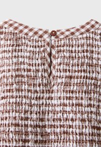 Tissu à carreaux marron et blanc avec texture smockée, avec un petit bouton marron et une fermeture par boucle en haut, au centre.