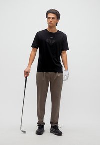 Νέος άνδρας με μαύρο μπλουζάκι και καφέ παντελόνι κρατώντας ένα μπαστούνι γκολφ, φορώντας λευκό γάντι γκολφ στο αριστερό του χέρι, στέκεται μπροστά σε απλό φόντο.