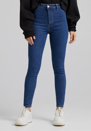Femme portant un jean skinny taille haute bleu, un haut à manches longues noir et des baskets blanches, debout devant un fond uni.