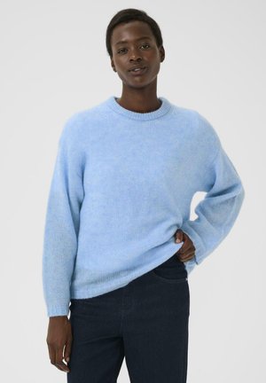 Vrouw in een lichtblauwe gebreide trui en donkere broek, staande met één hand op haar heup tegen een eenvoudige witte achtergrond.
