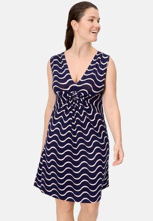 Mujer de cabello oscuro recogido en una coleta, vestida con un vestido sin mangas azul marino con rayas onduladas blancas y escote en V, sonriendo y mirando hacia su derecha.