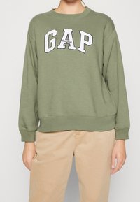 Felpa verde oliva con polsini e orlo a coste, con logo "GAP" ricamato in bianco. Indossata sopra pantaloni beige chiaro.