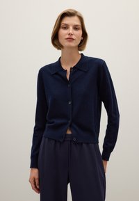 Marineblauwe cardigan van zacht materiaal, met een kraaghalslijn, knopenfront en geribde mouwen, gecombineerd met donkere, elastische taillebroeken.