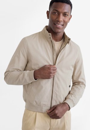 Uśmiechnięty mężczyzna zapinający beżową lekką kurtkę, ubrany w beżowe spodnie, stojący na tle białego, gładkiego tła.