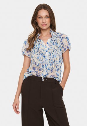 Blommig blus i genomskinligt tyg med blåa och lila blommor, volangkrage, korta ärmar och knäppning fram, ihop med mörka byxor.