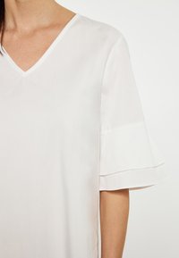 Weißes Blusenshirt mit V-Ausschnitt und kurzen, ausgestellten Ärmeln. Glattes Material mit leicht glänzender Textur. Keine sichtbaren Muster oder Akzente.