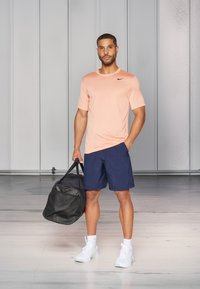 Athletic outfit featuring a light peach Nike t-shirt, navy shorts, and white sneakers, with a black duffel bag in hand.