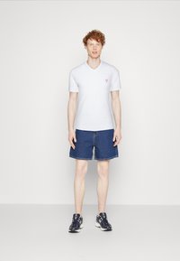 Hvid V-hals T-shirt med rødt logo, parret med blå denimshorts og sorte sportssko, med en neutral baggrund.