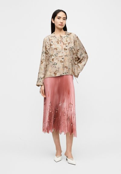 Κρεμ μπλούζα με floral μοτίβο και δετό λαιμόκοψη, συνδυασμένη με ροζ φούστα με πτυχές που διαθέτει κεντήματα με λουλούδια και κυματιστό τελείωμα, φορεμένη με λευκές γόβες.