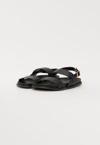 Sandalias de cuero negro con tiras entrelazadas, punta abierta, suela plana y un detalle de hebilla en el costado. Diseño minimalista con textura suave.