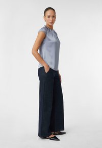 Helles blaues Satinbluse mit Rüschensäumen, kombiniert mit weit geschnittenen dunklen Denim-Hosen. Das Modell steht mit den Händen in den Taschen.