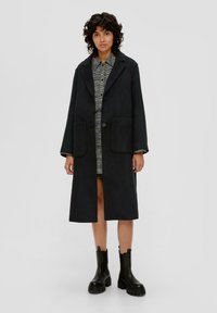 Schwarzer Wollmischungs-Langmantel mit Reverskragen, zwei vorderen Taschen und einem Knopfverschluss. Getragen über einem karierten Kleid und schwarzen Stiefeln.