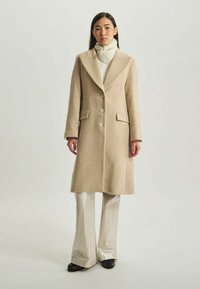 Manteau en laine beige avec de grands revers, boutons simple boutonnage et poches avant, porté sur un col roulé blanc et un pantalon clair brillant.