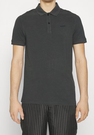 Mand iført en mørkegrå poloshirt med korte ærmer og to knapper, kombineret med sorte bukser med nålestriber, stående foran en hvid baggrund.