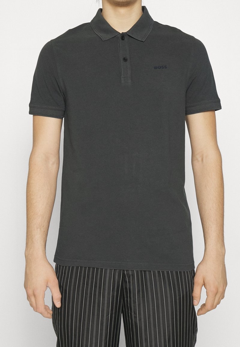 Homme portant un polo manches courtes gris foncé à deux boutons, associé à un pantalon noir à fines rayures, debout devant un fond blanc.