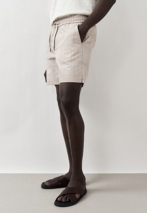 Férfi bézs kockás rövidnadrágban és barna bőr szandálban, áll egy egyszínű világos háttér előtt, egyik kezét a zsebében tartva.