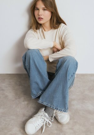 Suéter beige con adornos en los hombros, jeans azules de pierna ancha y zapatillas blancas. Posición sentado contra un fondo claro.